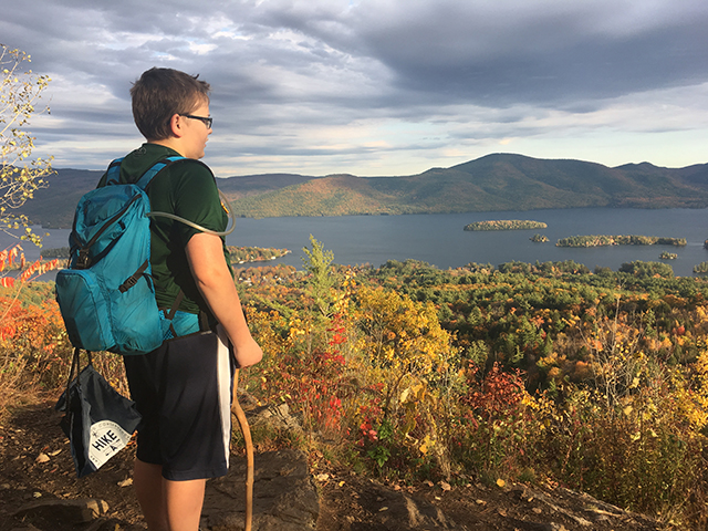 A boy with a backpack stand at top of Bradley's Point staring towards Lake George.