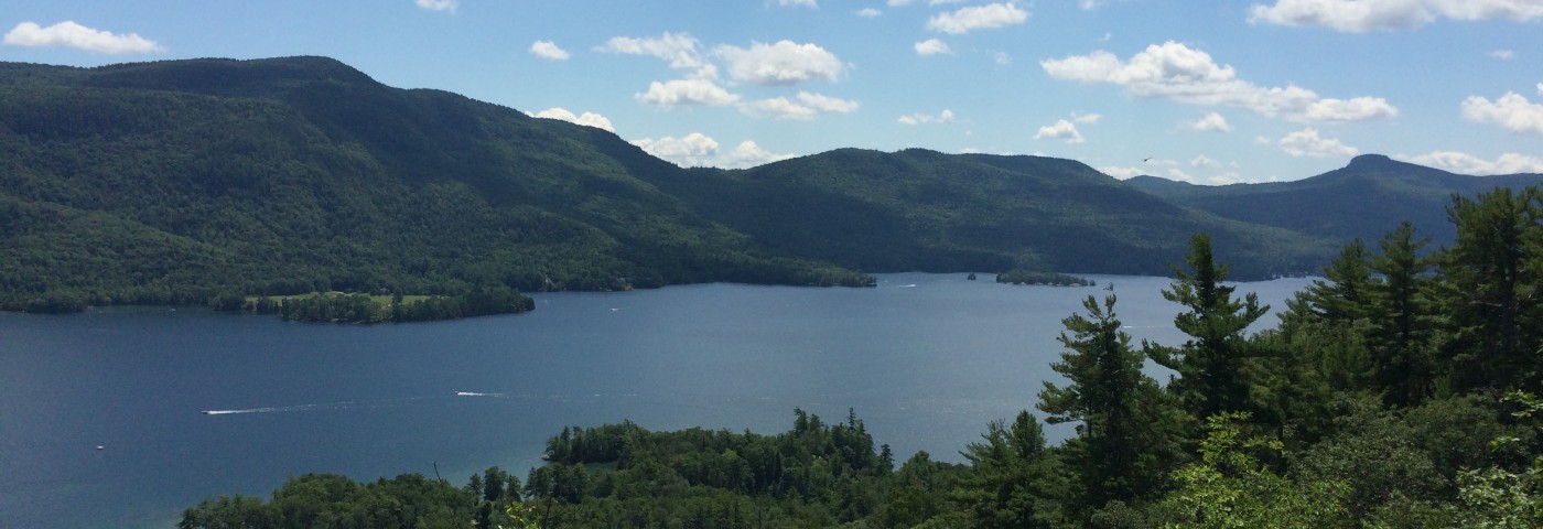 Panoramic view of Terzian Woodlot with a serene lake framed by mountains, trees, and a bright blue sky.