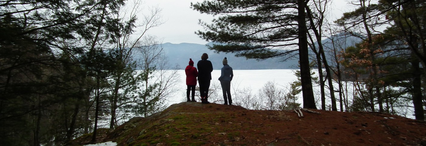 Three people stand at the edge of a hill over looking Lake George with visible mountains in the background.