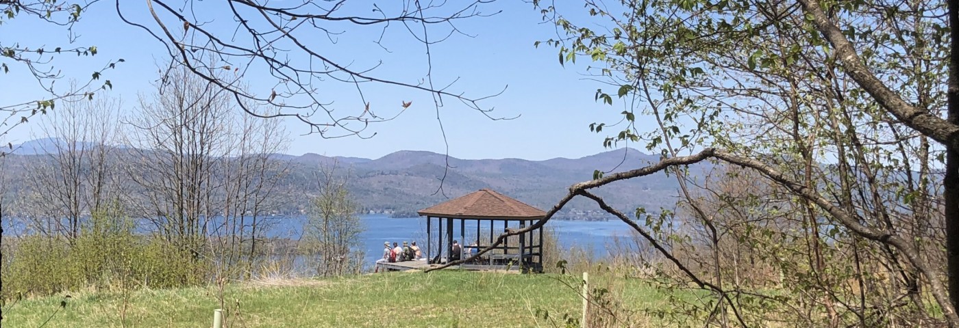 Several people sit by a wooden gazebo looking towards a lake and mountains.