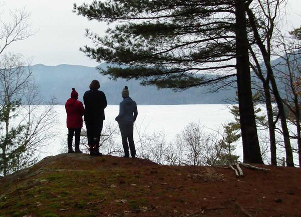 Three people stand at the edge of a hill over looking Lake George with visible mountains in the background.