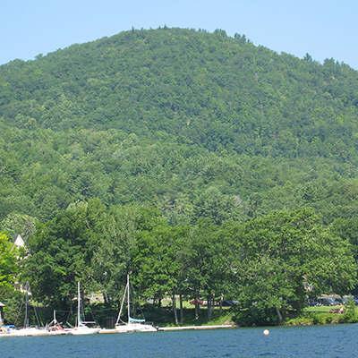 Boats docked by the shore of a tall mountain covered it bright green trees.