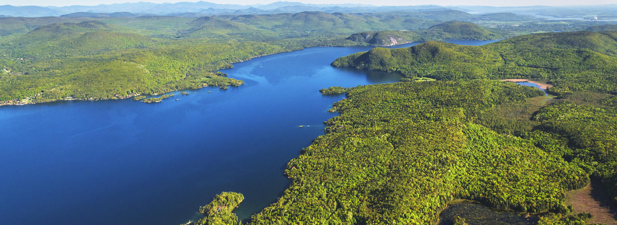 An aerial view of a lake surrounded by trees and mountains. The lake is a deep blue color, and the trees are a mix of green and yellow.