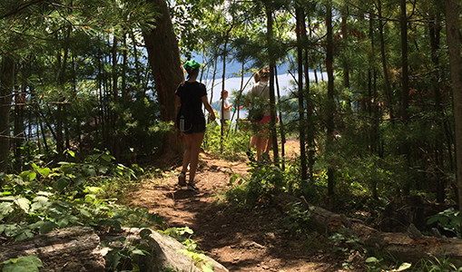 Three people walking through a winding path towards lake George. The path is surrounded by lush green trees.