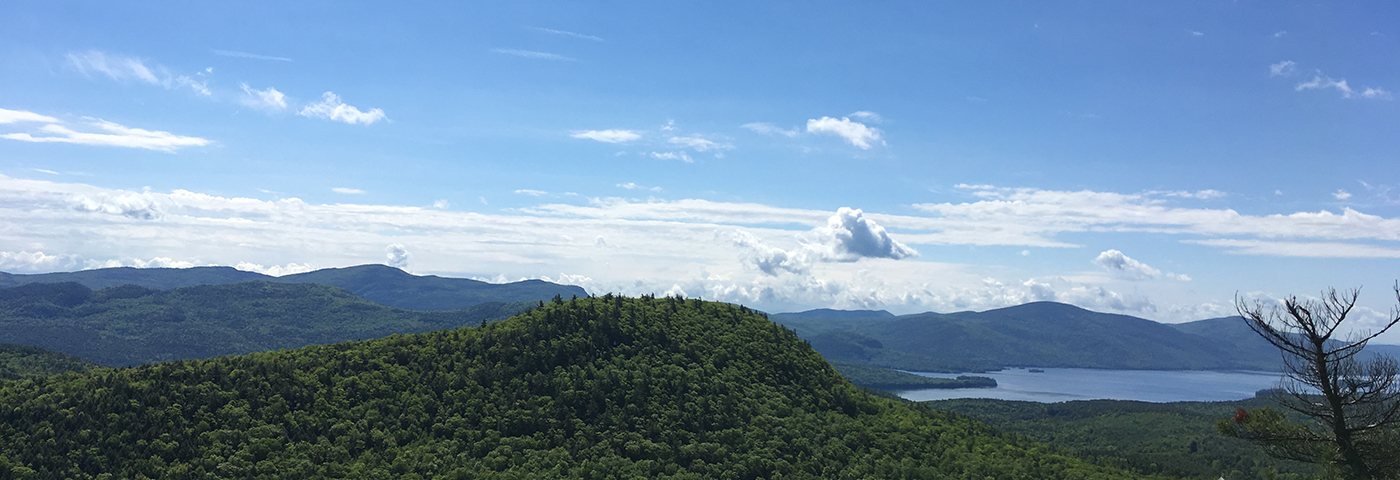 A panaromic view of Pole Hill with Lake George in the distance and several mountains in the background.