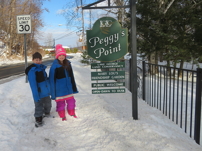Two children stand in a snowy road next to the LGLC Peggy's Point sign. Below Peggy's Point sign there are several other signs that read