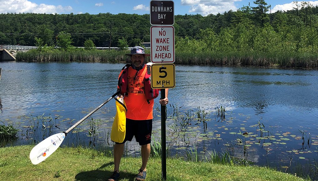 A man in an orange shirt with a life vest and paddle stands next to multiple signs that read: (from top to bottom) Bunhams Bay, No Wake Zone Ahead, and 5 PMH. Behind him is water with some lily pads and a dense forest in the background.