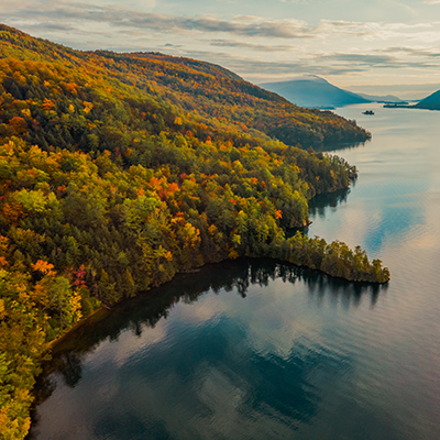 An aerial view of a lake surrounded by trees in the fall. The trees are ablaze with red, orange, and yellow leaves, contrasting with the blue water of the lake.