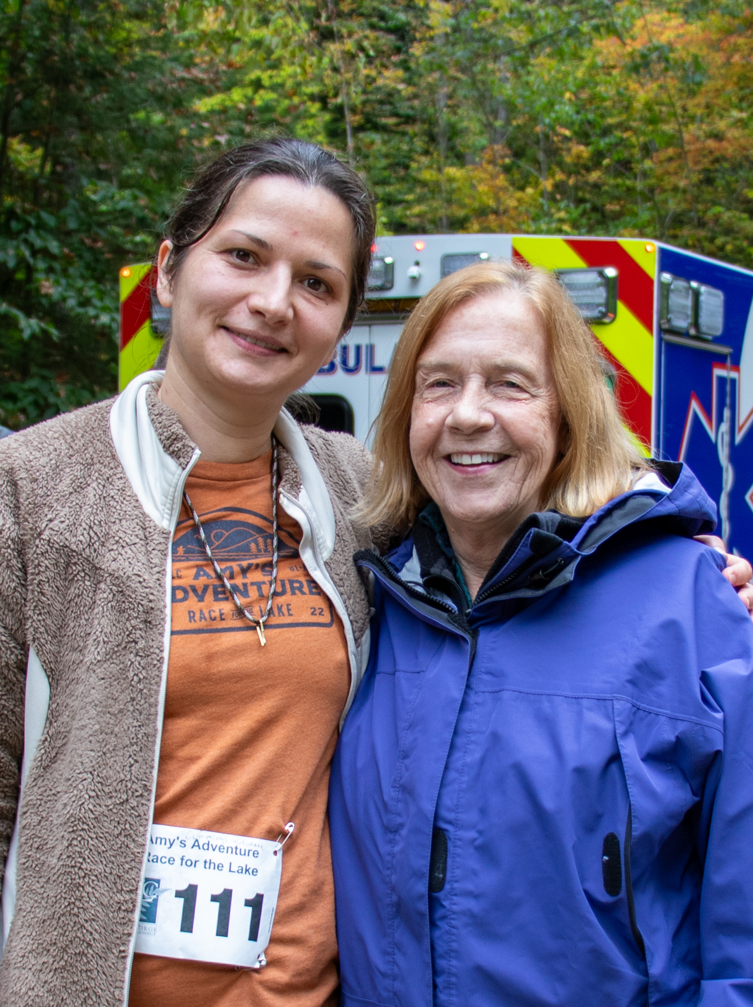 Two women close to each other pose smiling in front of an ambulance.