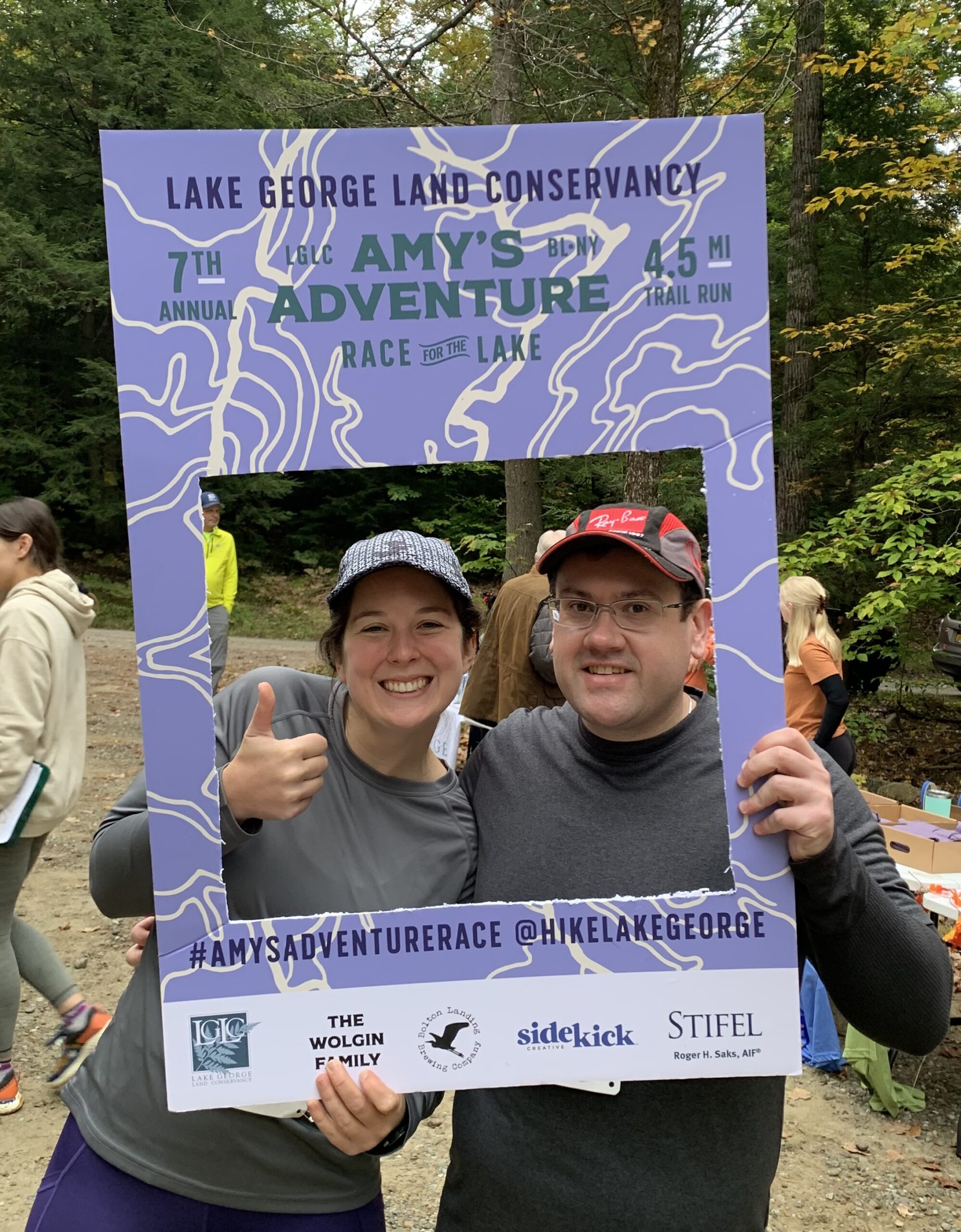 A couple posing with a sign for the Lake George Land Conservancy featuring Amy's Adventure Race for The Lake.