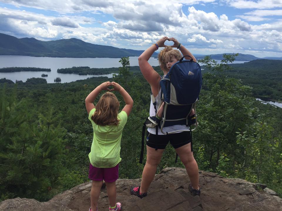 A mother with a baby in a back carrier stands next to her daughter on the summit of The Pinnacle Preserve. She and her daughter are making heart shapes with their hands, held over their heads, with Lake George in the background.