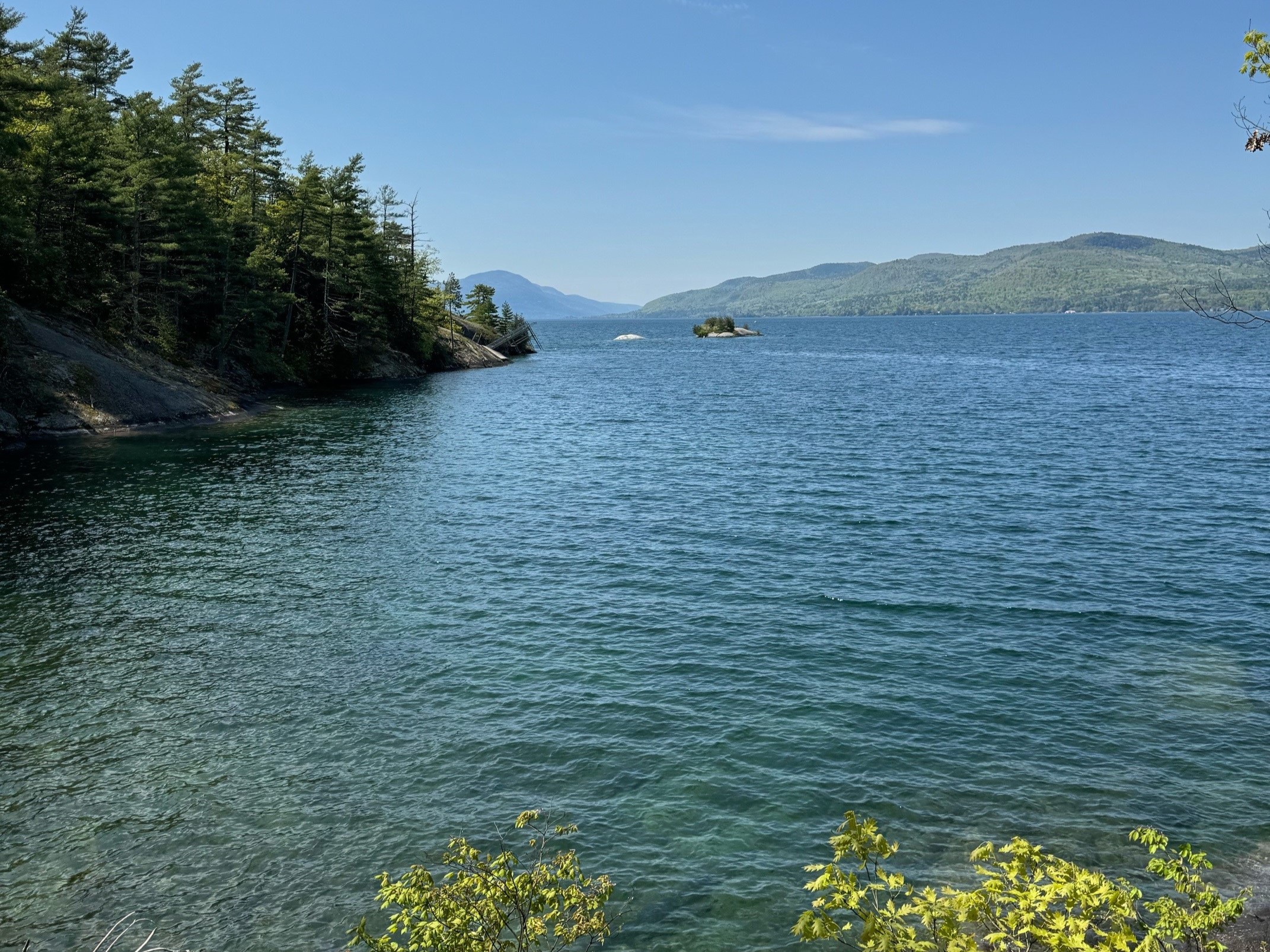 Clear blue water of Lake George ripples in the wind. Green mountains frame the backdrop, with a forested shoreline in the forefront.