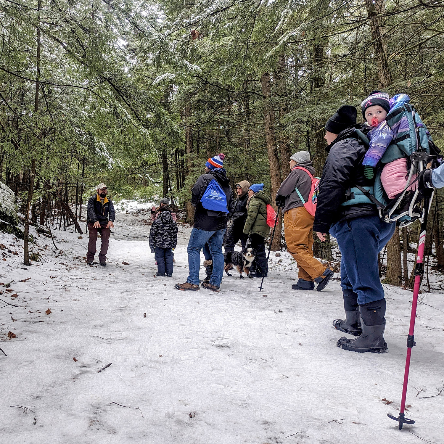 A group of adults and children stand on a snowy wooded trail.