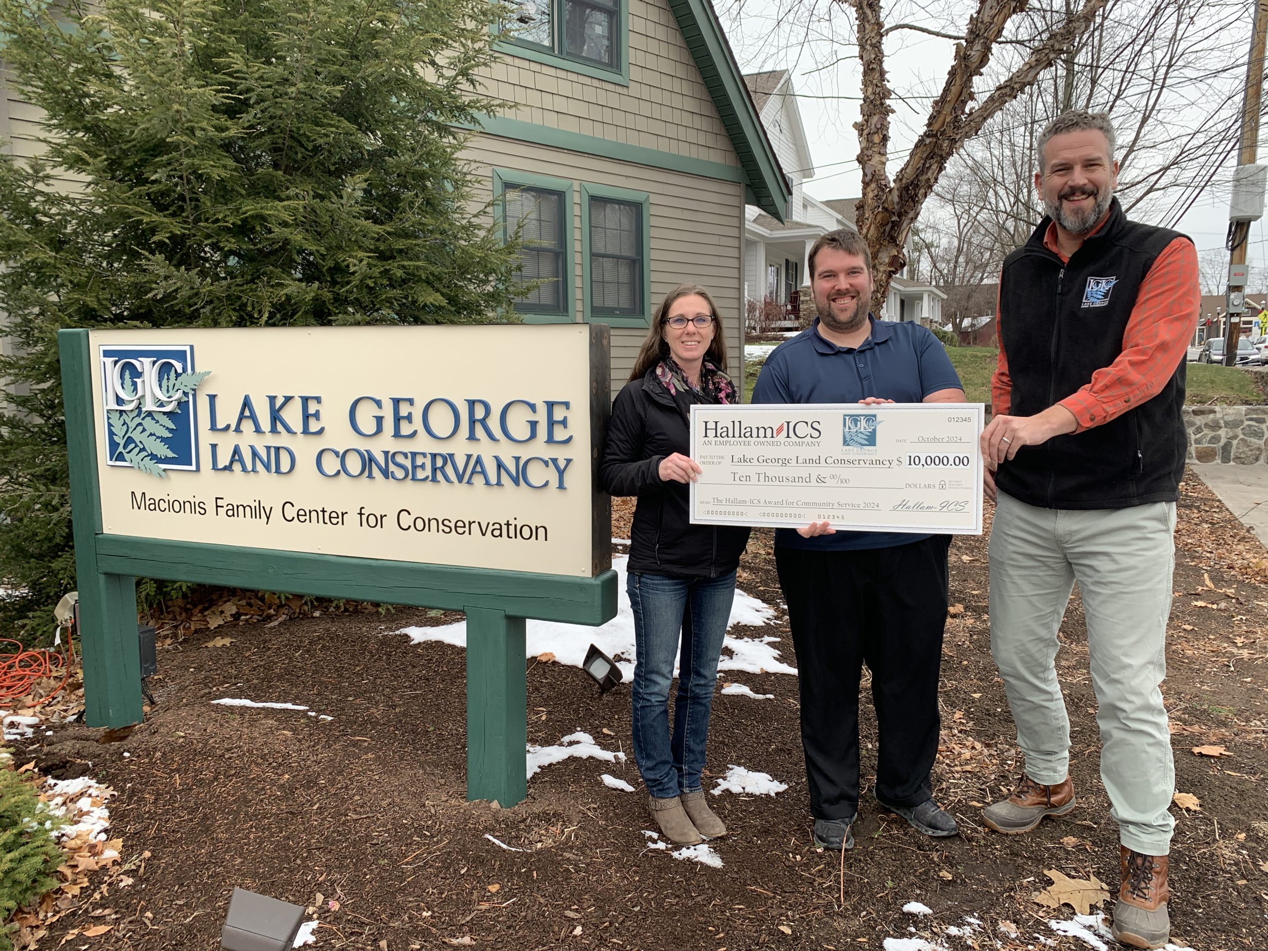 Hallam-ICS employees Julie Montenegro and Jason Barry stand next to LGLC Executive Director Mike Horn by a sign showing the LGLC logo. The three hold a large symbolic check.