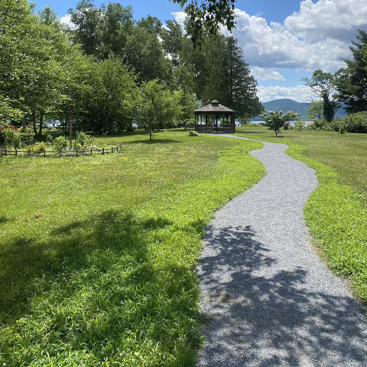 Green grass lawn extends on either side of a compact gravel path. A small gazebo is at the end of the path.